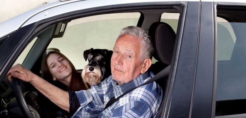 Smiling senior driver with a dog and younger passenger in the car, representing how auto insurance age brackets can span multiple generations of drivers.