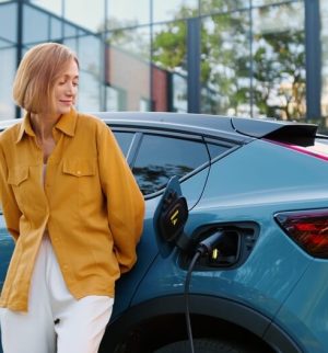 Woman standing beside a charging electric car outside an office building, highlighting electric vehicle insurance